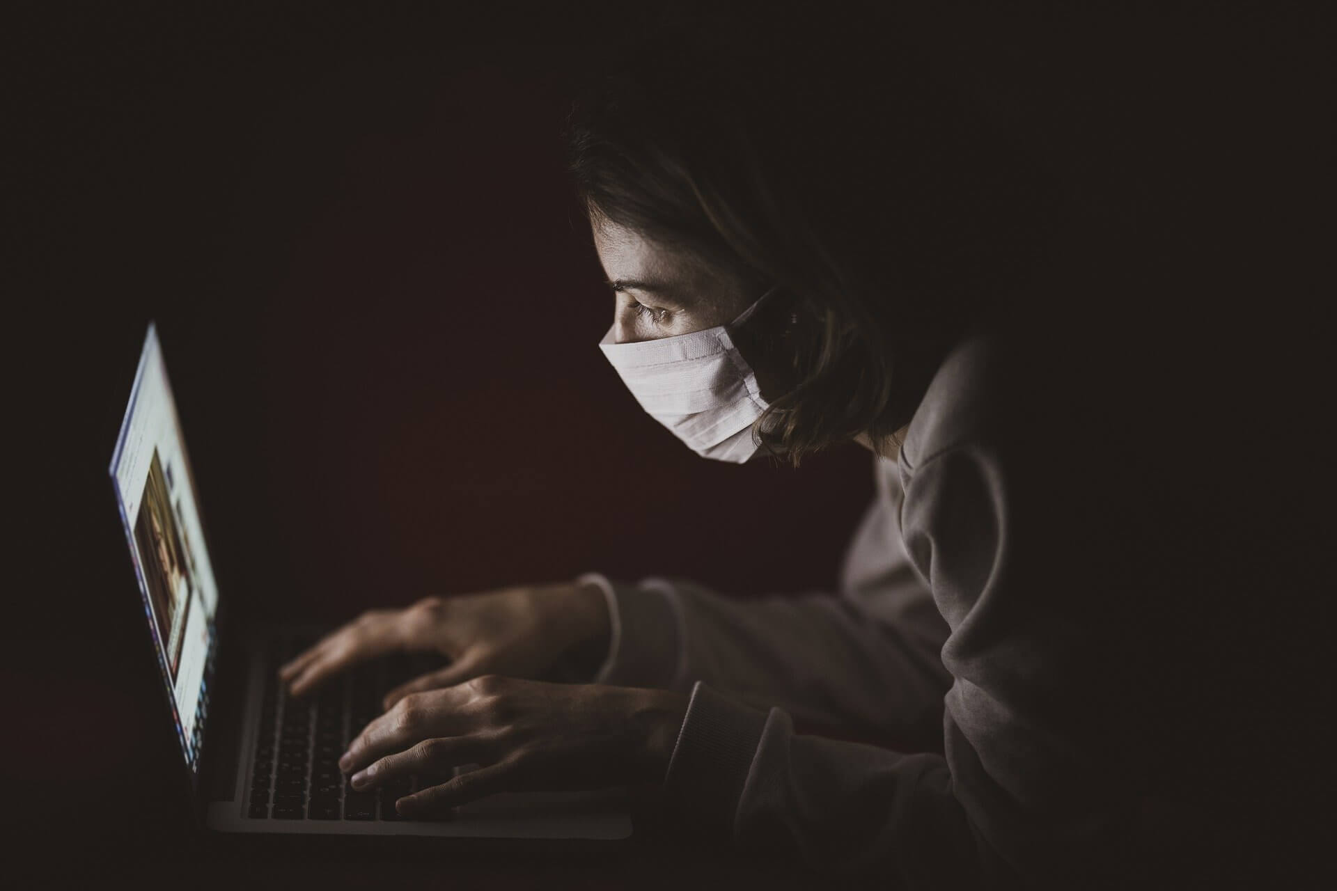 person typing on a laptop while sitting in the dark wearing a surgical mask like those worn during COVID-19 pandemic